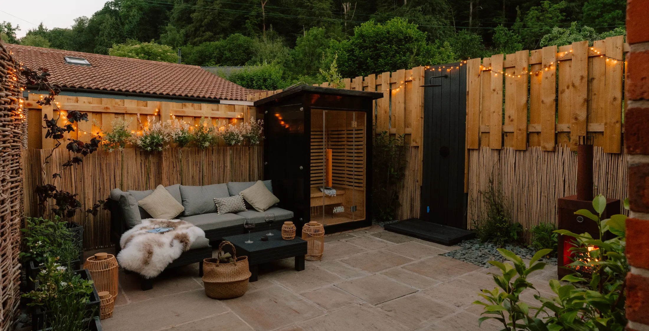 Luxury stone cottage courtyard terrace at night featuring an lit infrared wooden sauna for two, an outdoor shower, comfortable outdoor sofas, a wood-fired pizza oven, and a glowing patio heater.