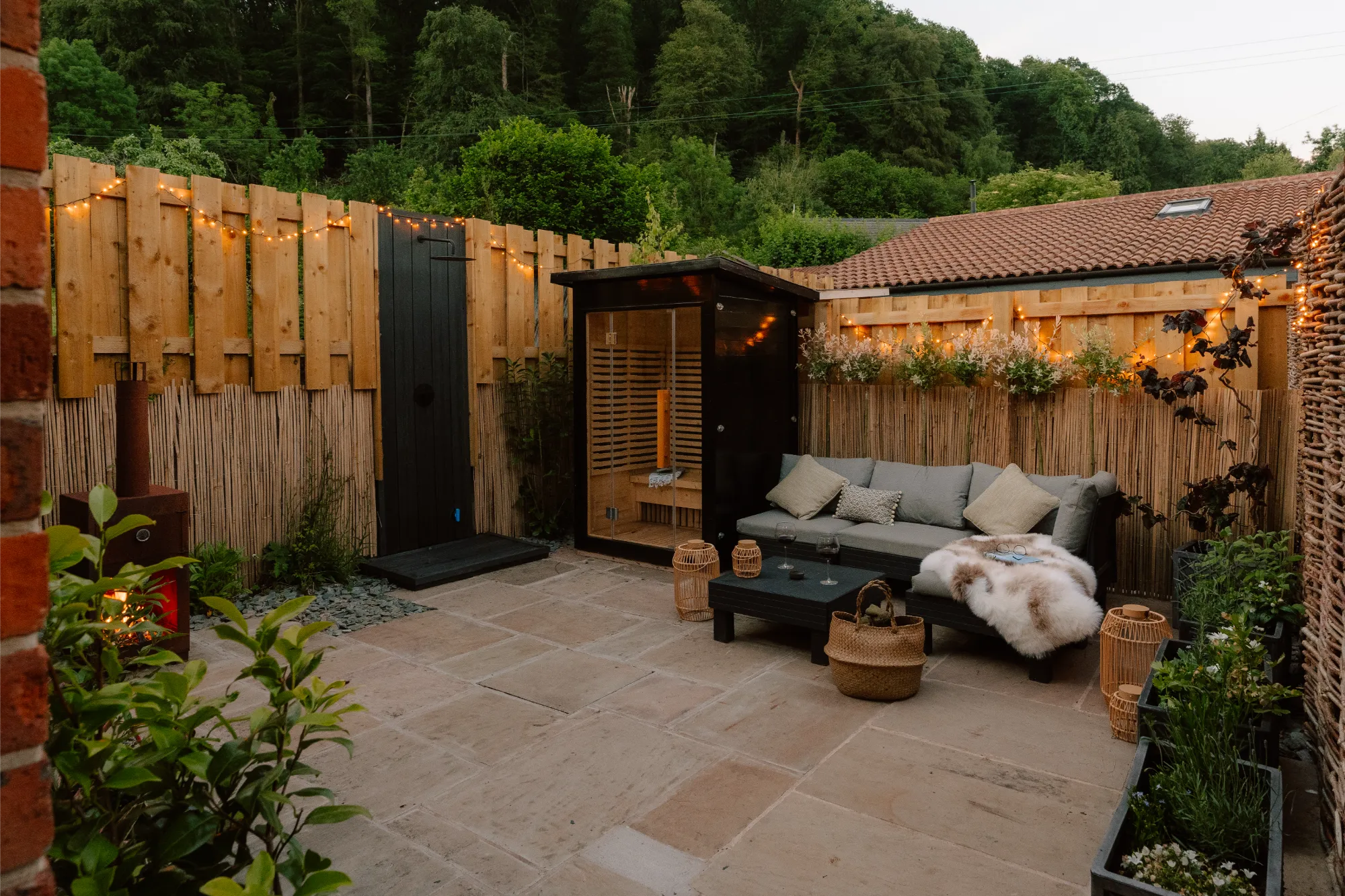 Luxury stone cottage courtyard terrace at night featuring an lit infrared wooden sauna for two, an outdoor shower, comfortable outdoor sofas, a wood-fired pizza oven, and a glowing patio heater.