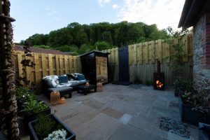 Luxury stone cottage courtyard terrace at night featuring an lit infrared wooden sauna for two, an outdoor shower, comfortable outdoor sofas, a wood-fired pizza oven, and a glowing patio heater.